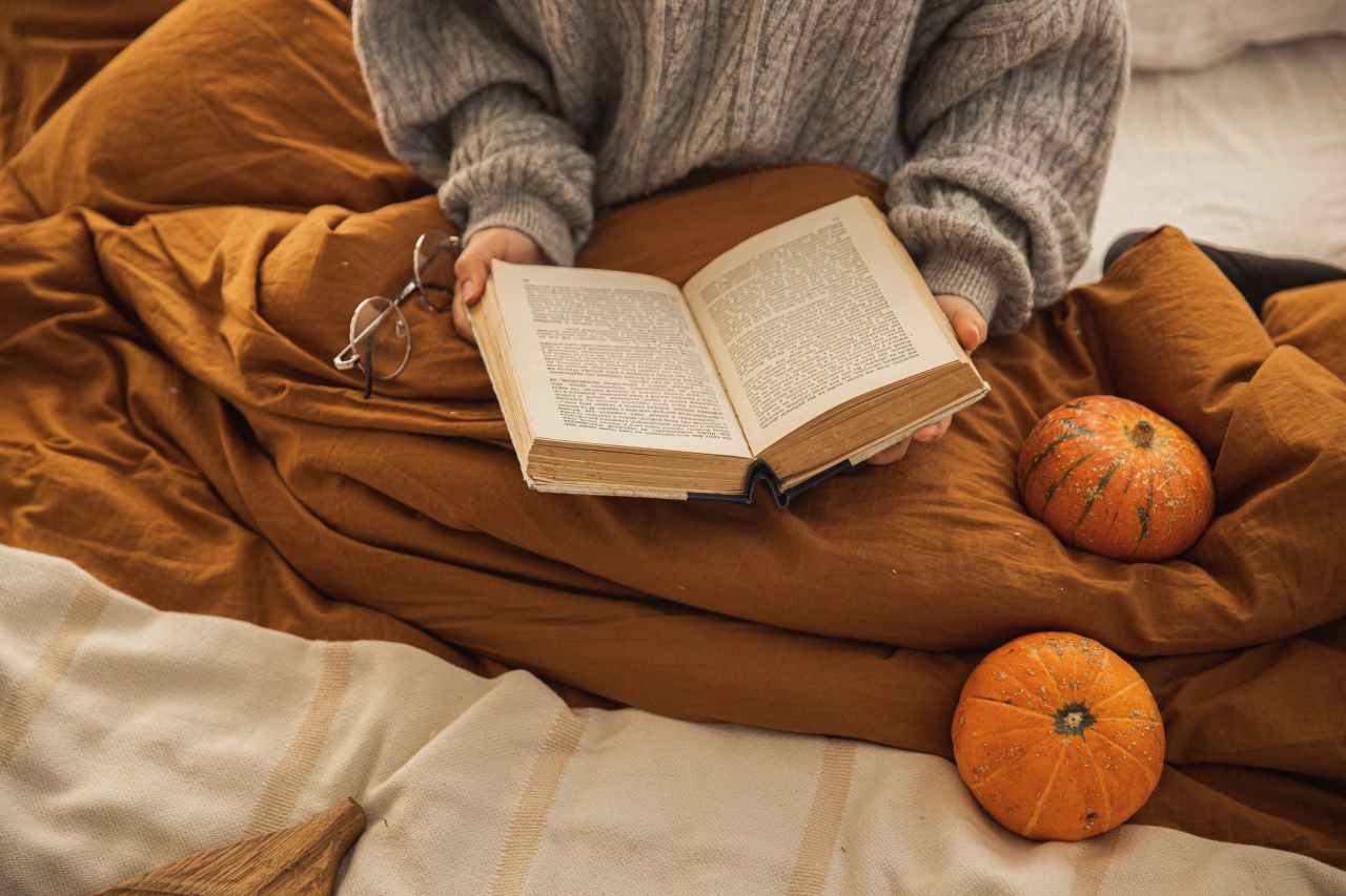 Someone reads a book in bed, with 2 pumpkins beside them.