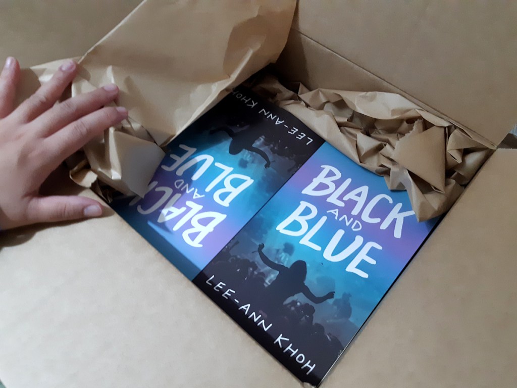 Hand opens a box containing copies of Black and Blue by Lee-Ann Khoh.