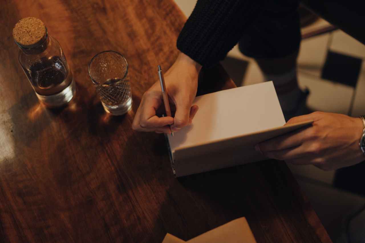 Person signs a book on a wooden table with a glass and corked bottle on it.