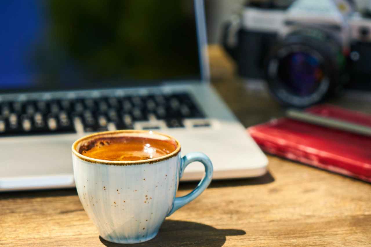 A cup of coffee on a wooden desk with a laptop, camera, and notebook.