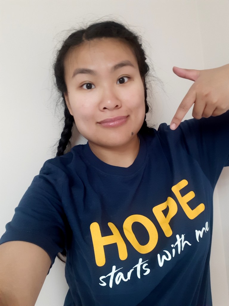 Lee-Ann, a woman with long black hair tied into two French braids, points to the text on her dark blue T-shirt that reads "Hope starts with me".