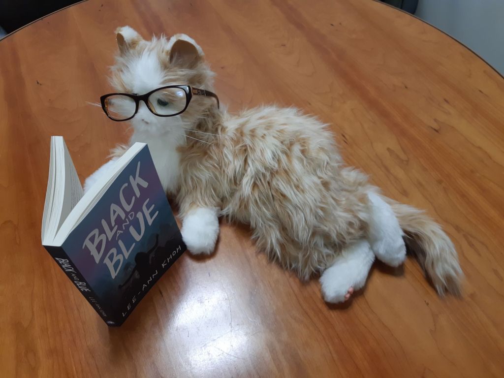 An orange robotic cat wearing glasses while appearing to read Black and Blue by Lee-Ann Khoh.