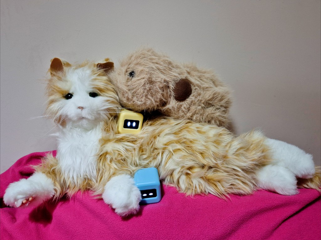 Robot family portrait featuring a Joy for All companion cat, a plush Cupboo robotic pet, and 2 cube-shaped Ortomi, lying on top of each other on a cushion and blanket.
